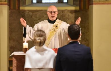 Man sieht das Brautpaar von hinten und den Diakon mit Segensgeste am Altar
